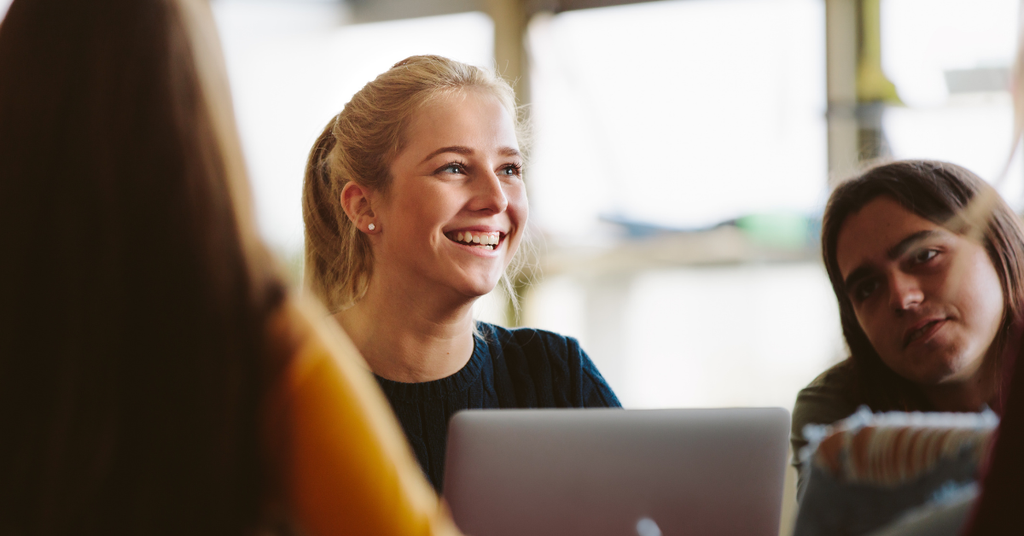 A college student working on a laptop with her classmates smiles into the distance.