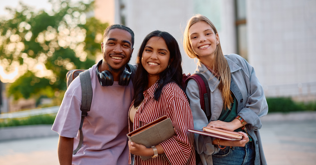 A group of three college students stand outside on a sunny day. They lean into one another, smiling happily at the camera.