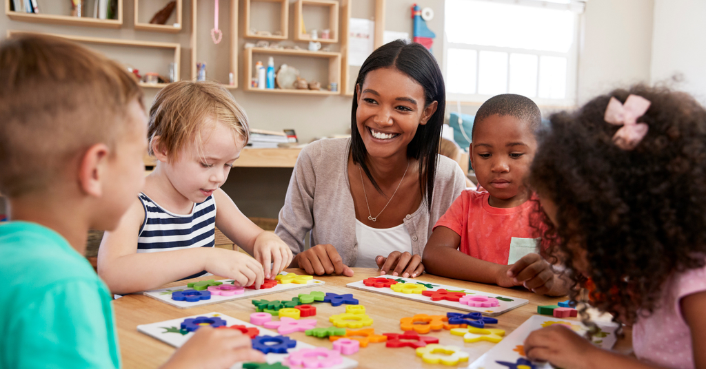 An Early Childhood Educator smiles happily while sitting at a table with young children working on puzzles.